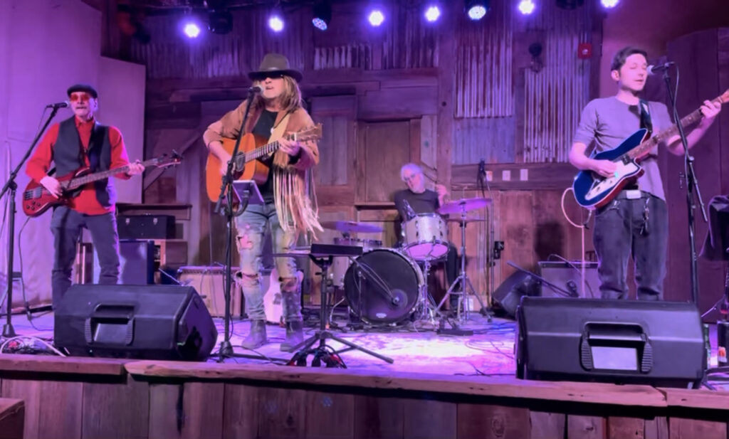 Sugar mountain - Visit Angel Fire New Mexico Band performing on a wooden stage with bass, guitar, drums, and microphones under purple stage lights.