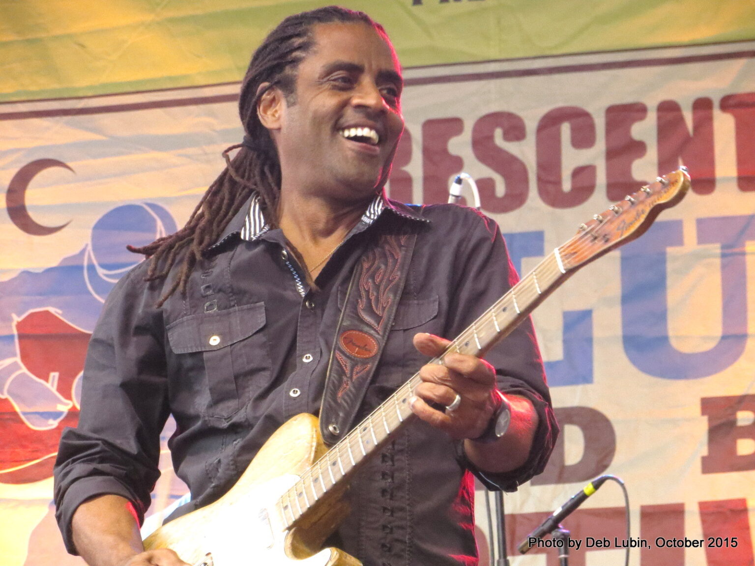 Male guitarist with dreadlocks playing a light-colored electric guitar on stage, smiling toward the crowd, with a banner in the background.