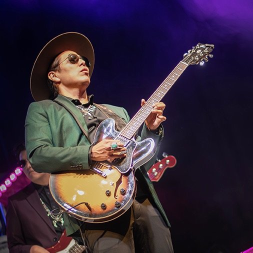 Male guitarist wearing a wide-brimmed hat and sunglasses, playing a sunburst electric guitar on stage.