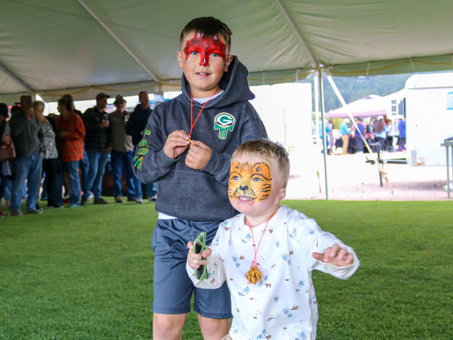 Two smiling boys with face paint under a canopy at an outdoor festival; one has red dragon makeup, the other tiger makeup nearby.