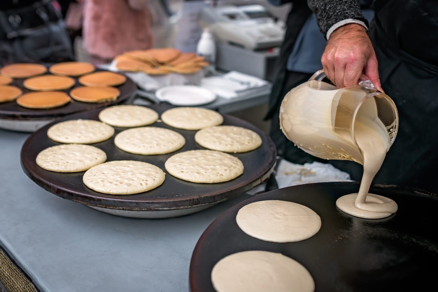 Pancake batter being poured onto round griddles at a street food stall, several pancakes already cooking nearby