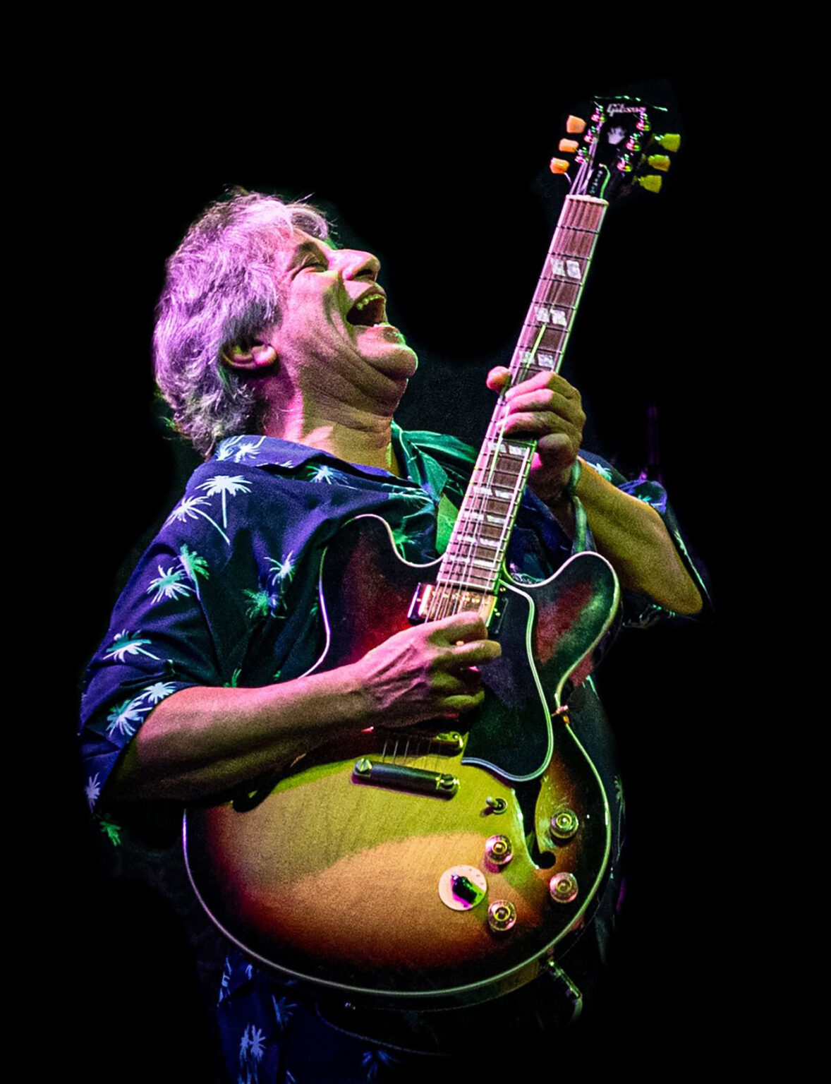 Guitarist wearing a palm-tree shirt passionately playing a sunburst electric guitar on a dark stage with colorful lights.