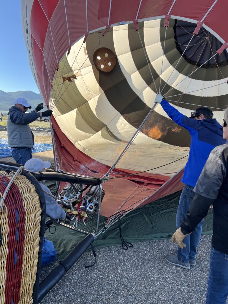 Crew members hold ropes as a red and cream hot air balloon is inflated near the wicker basket with burners visible in the foreground.