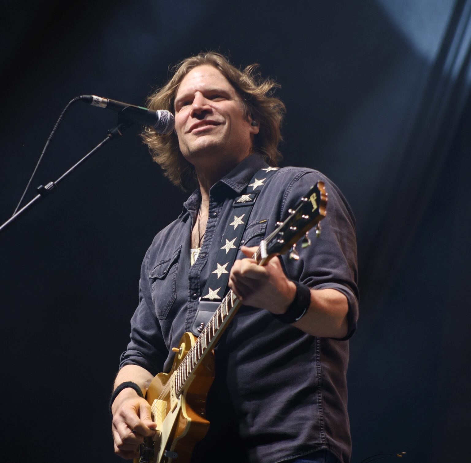 Male guitarist performing on stage with a microphone nearby, smiling toward the audience.