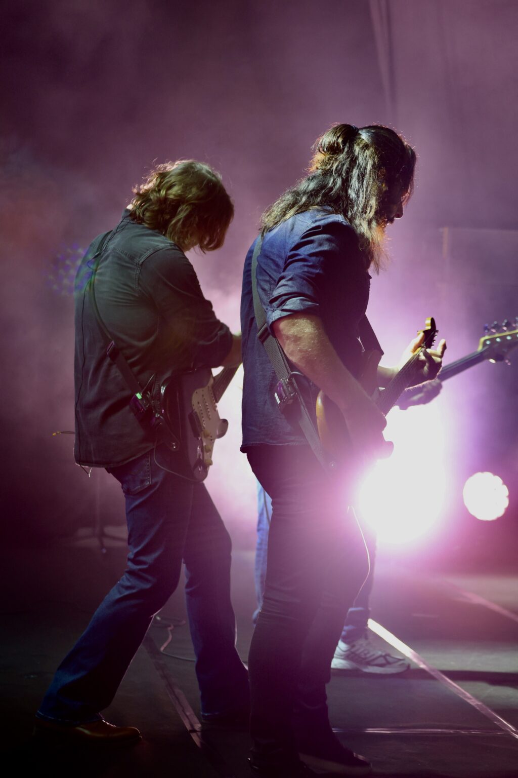 Two guitarists performing on a smoky stage with pink and purple backlighting.