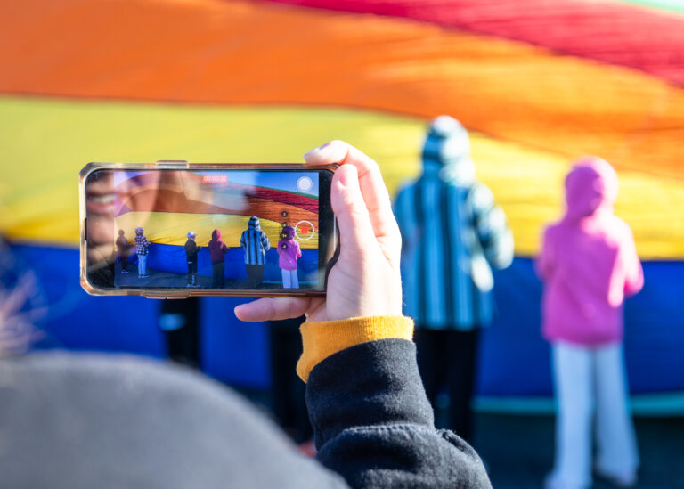 Hand holding a smartphone filming a group of kids in front of a large colorful inflatable slide in the background.