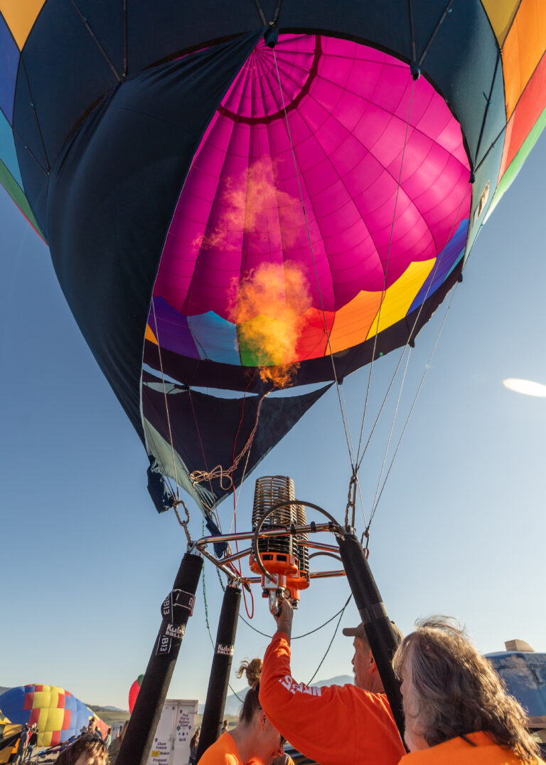 Close-up of a colorful hot air balloon being prepared for flight, with burners firing and crew in orange shirts nearby.
