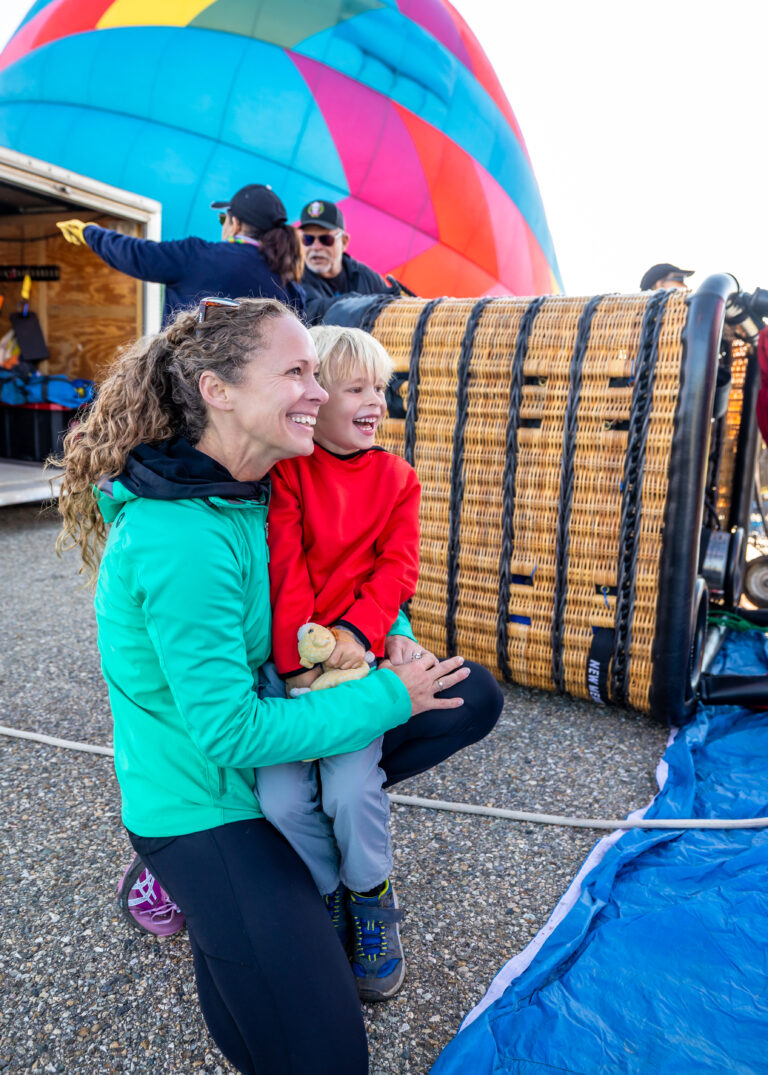 Smiling woman in a teal jacket holds a laughing young boy in a red shirt, with a colorful hot air balloon and wicker basket behind them.