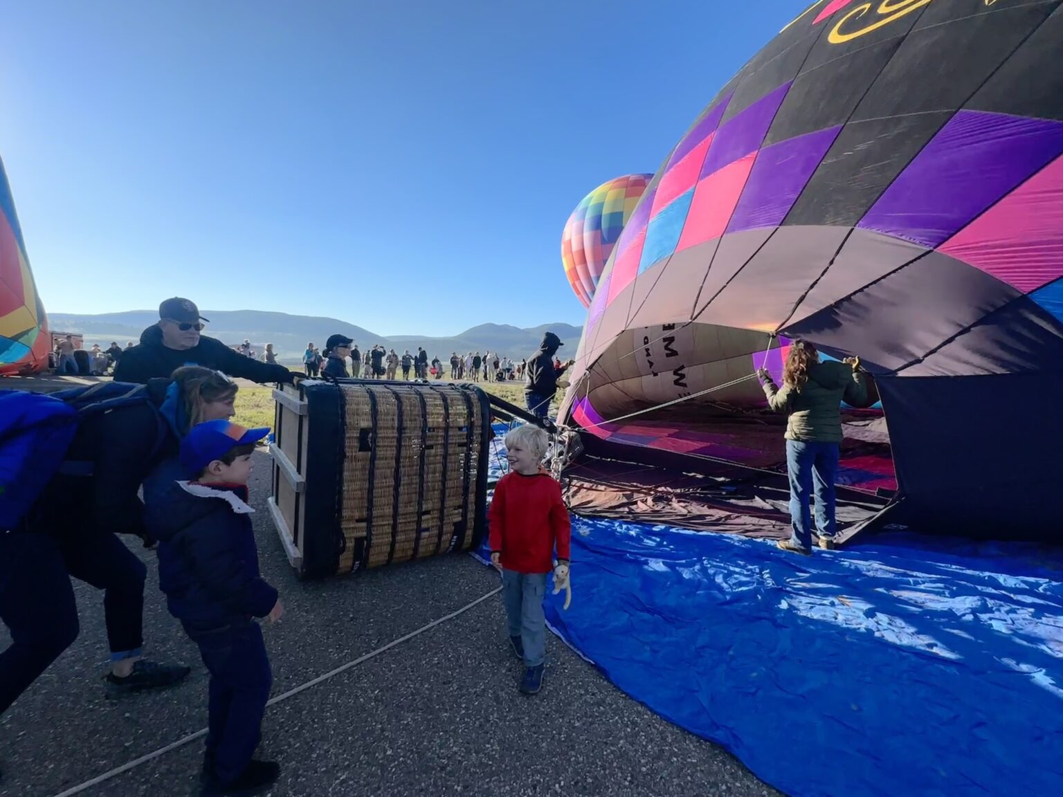 People preparing colorful hot air balloons for launch on an open field, baskets and a blue tarp visible in the foreground.