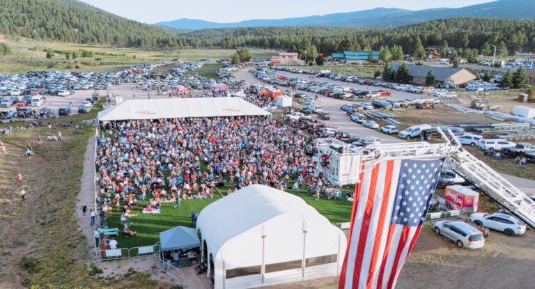 Mountain View Event Center - Visit Angel Fire New Mexico Huge outdoor festival crowd gathered under a white event tent, with a tall American flag in the foreground and mountains in the distance.