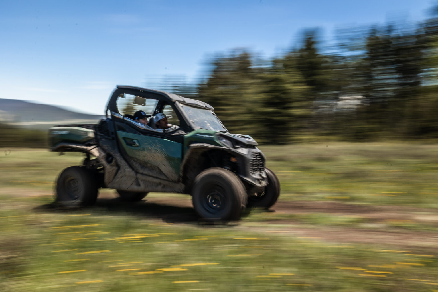 Two riders in a teal side-by-side UTV speeding through a grassy field, motion blur in the background.