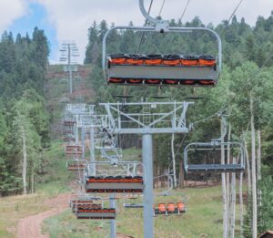 Empty ski lift chair approaching over a forested mountain slope. Terminology: chair with orange seats hanging from a cable.