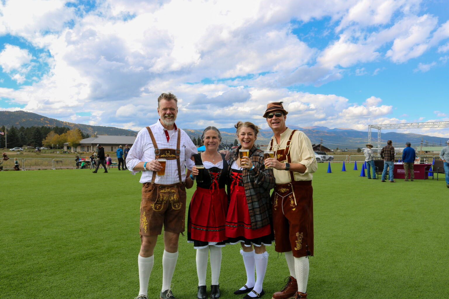 Four adults in traditional Bavarian outfits (lederhosen and dirndl) posing with beers on a grassy field at an outdoor festival under a blue sky.