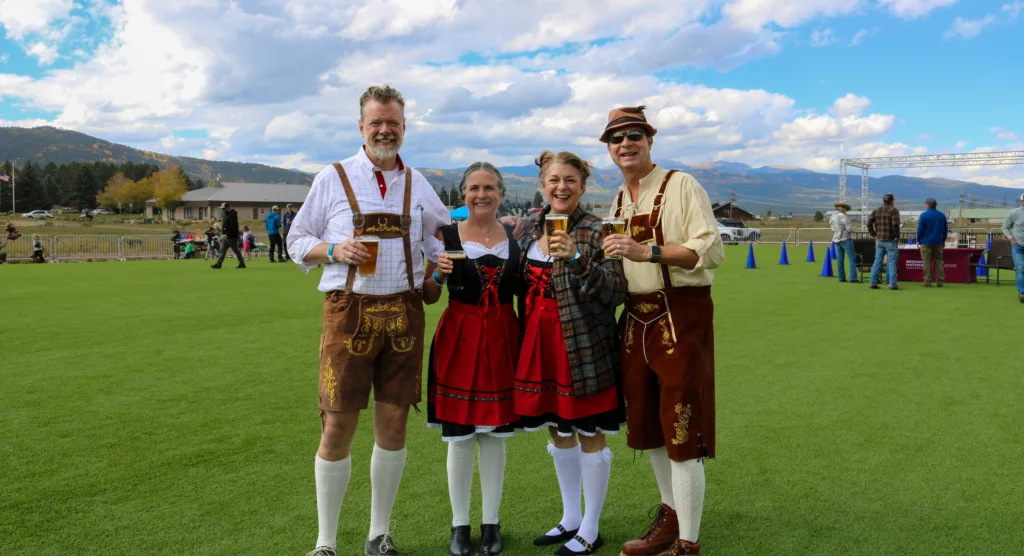 Four adults in traditional Bavarian outfits (lederhosen and dirndl) posing with beers on a grassy field at an outdoor festival under a blue sky.