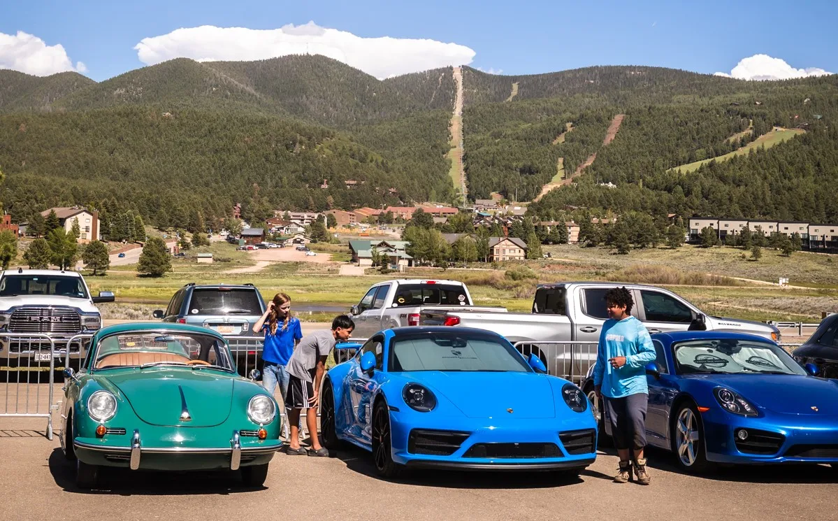 Group of people admiring a row of sports cars parked in a lot with mountains in the background; turquoise vintage car, blue Porsche 911, and other blue cars visible.