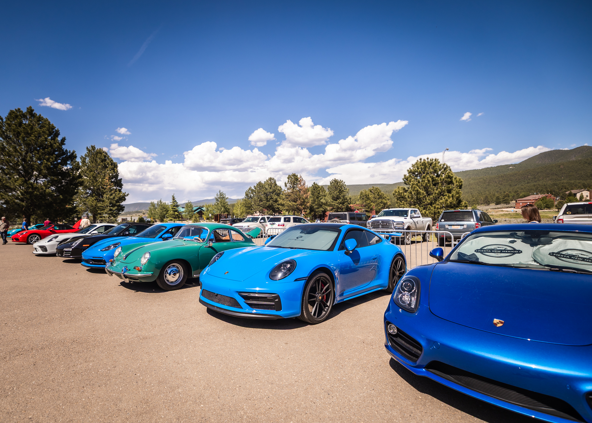 Row of blue sports cars parked in an outdoor car show with trees and hills in the background.