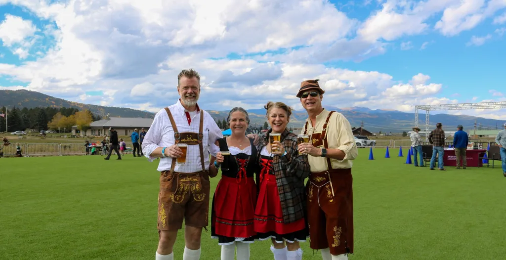 Four adults in Bavarian outfits pose with beer at an outdoor festival, mountains in the background, blue sky and clouds above. But concise alt text only.