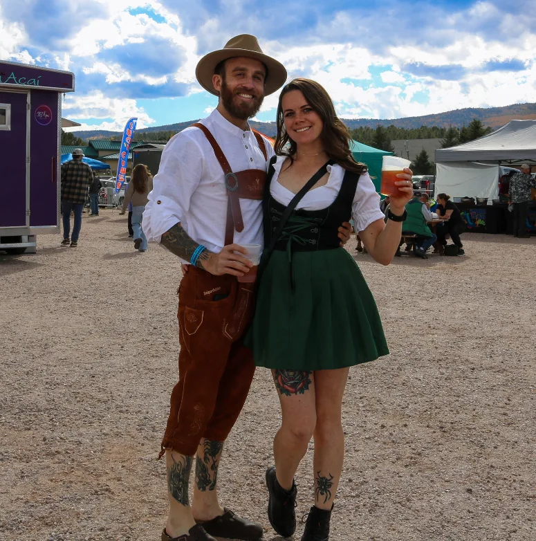 Smiling couple in lederhosen and a dirndl pose with beer at an outdoor festival, tents and vendors in the background.