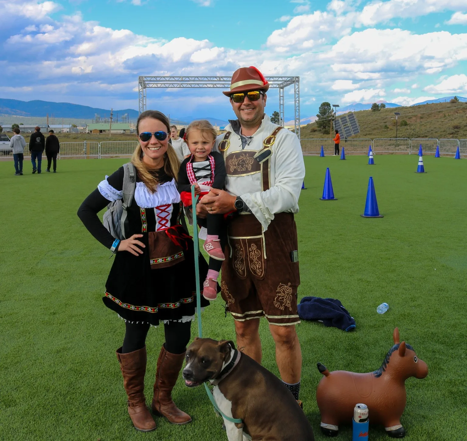Family in festive costumes poses on a grassy field with a dog at an outdoor event, man in lederhosen and woman in a dirndl with child on his arm